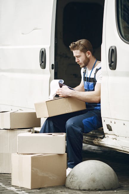 A man with a beard and short brown hair, wearing a striped T-shirt and blue overalls, is seated at the open side door of a white removal van. He appears to be reviewing a clipboard or moving paperwork