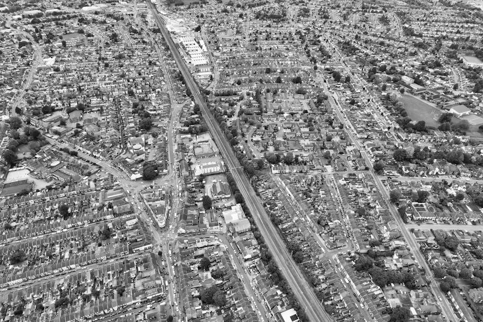 A black-and-white aerial photograph showing a densely populated residential area during house relocation. The image captures numerous tightly packed terraced houses with small front gardens, narrow streets, and a network of alleys. A railway line cuts through the scene, accompanied by a railway station visible in the upper part of the image. In the lower part, a moving van from Man with Van Welling is loading boxed household items, including cardboard boxes, plastic-wrapped furniture, and items on blankets. The van, parked on the pavement outside one of the homes, has its side door open with movers seen lifting and transporting boxes and furniture, preparing for a home relocation service in Bellegrove Road to Welling Station area. The surroundings include trees, driveways, and adjacent streets, with adequate space for the furniture transport and equipment used during the moving process, such as trolleys and straps, visible in the scene.