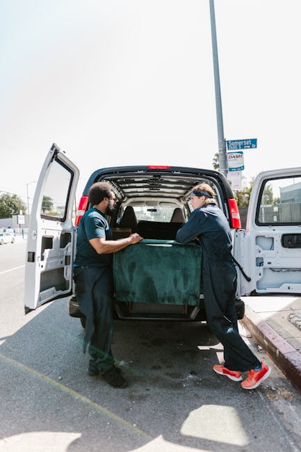 Two professional movers from Man with Van Welling are loading a large, green upholstered piece of furniture into the back of a white panel van parked on a street in Welling. The interior of the van has an open rear door and features protective padding along the ceiling, indicating a careful packing and moving process. One mover, wearing glasses and a dark jacket, is guiding the furniture inside while the other, dressed in a blue shirt, helps lift and position it. The scene takes place on a bright day, with a street sign visible in the background and urban surroundings including a sidewalk curb and parked vehicles. The image captures the key elements of home relocation, furniture transport, and packing of household items, reflecting the logistics involved in local removals services offered by Man with Van Welling.