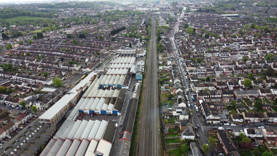 A black-and-white aerial photograph showing a densely populated residential area during house relocation. The image captures numerous tightly packed terraced houses with small front gardens, narrow streets, and a network of alleys. A railway line cuts through the scene, accompanied by a railway station visible in the upper part of the image. In the lower part, a moving van from Man with Van Welling is loading boxed household items, including cardboard boxes, plastic-wrapped furniture, and items on blankets. The van, parked on the pavement outside one of the homes, has its side door open with movers seen lifting and transporting boxes and furniture, preparing for a home relocation service in Bellegrove Road to Welling Station area. The surroundings include trees, driveways, and adjacent streets, with adequate space for the furniture transport and equipment used during the moving process, such as trolleys and straps, visible in the scene.