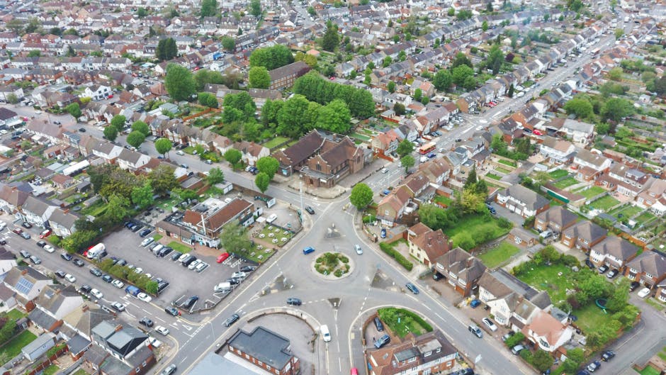 An aerial view of a residential neighbourhood showing a roundabout with several cars navigating the intersection, surrounded by numerous houses with varied roofing styles, gardens, and driveway parking. The image captures a section of the street corner, with some vehicles parked along the pavements and in on-site parking lots. Trees and shrubbery are visible throughout the area, providing greenery amid the housing. The natural daylight highlights the different textures of the rooftops and the layout of the homes. This scene illustrates a typical suburban environment suitable for house removals and furniture transport logistics, as part of a comprehensive home relocation process. The photograph, associated with Man with Van Welling, reflects the typical urban setting where professional removal services may operate, including loading furniture and boxes into vans for transport to new addresses.