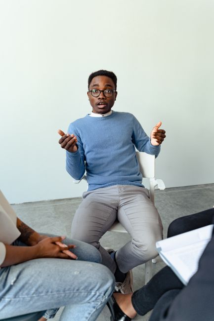 A man with glasses and wearing a blue sweater sits in a white chair inside a room with plain light-colored walls and a concrete floor. He is engaged in a discussion, with his hands gesturing expressive movements. Part of another person is visible in the foreground, sitting with crossed legs, wearing jeans, and holding a rolled-up paper or document. The setting suggests an informal consultation or briefing, possibly related to house removals or moving services such as packing or planning for a home relocation. There are no furniture or moving equipment visible in this specific image, but the context aligns with professional discussions about the details involved in furniture transport, loading, or obtaining permits from local authorities like Bexley Council for a full domestic move. The natural lighting and neutral background underscore a professional yet approachable environment for discussing relocation logistics, as facilitated by companies like Man with Van Welling.