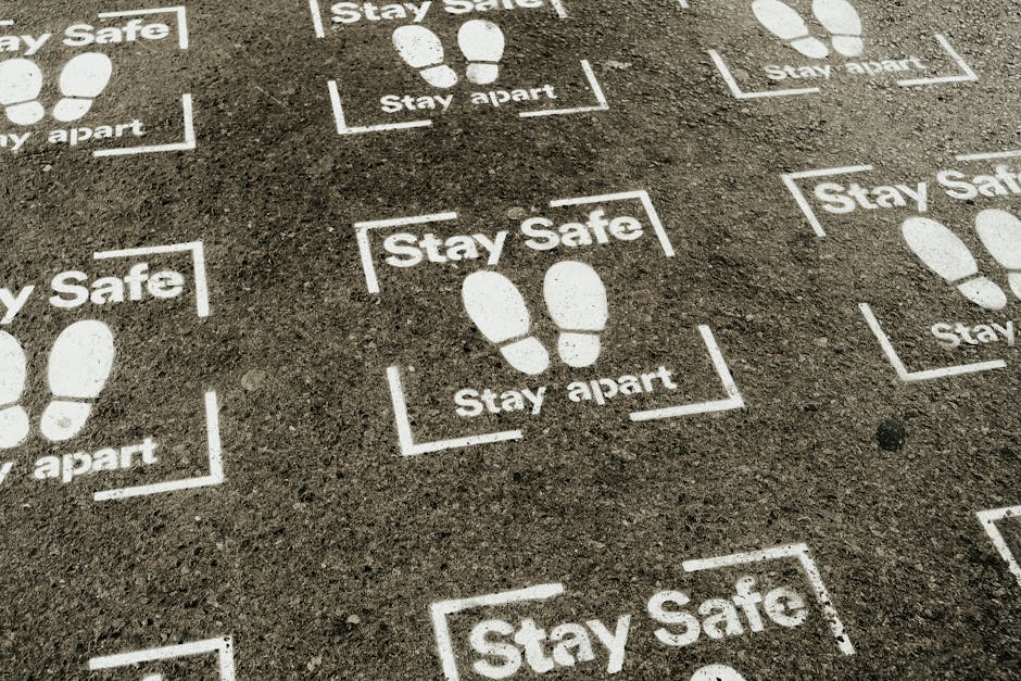 Close-up image of a paved ground surface with white stenciled markings and messages related to social distancing, including footprints inside rectangular borders and the words 'Stay Safe' and 'Stay apart.' The markings are evenly spaced on the asphalt, indicating designated safe areas for walking or queuing during a move or home relocation process. These markings are visible in an outdoor setting, possibly a driveway or parking area, with natural lighting highlighting the textured surface. Occasionally, the markings in the scene are associated with the logistical aspects of furniture transport and packing during a house move, as managed by [COMPANY_NAME], a professional removals service. The overall clean and maintained appearance of the surface suggests an organized approach to safe moving practices during a home relocation or delivery process.