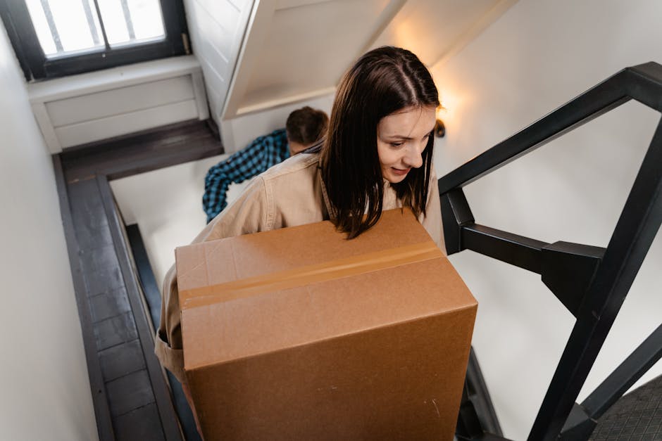 A woman with dark brown hair, wearing a beige coat, is carefully carrying a large cardboard box up a narrow staircase in a terraced home during a home relocation. Behind her, a man wearing a blue checkered shirt is also ascending the stairs, holding a similar box. The staircase features dark wooden steps with a black metal railing on the side, leading to an upper floor visible at the top. Natural light is coming through a window near the staircase, illuminating the scene. The concrete or tiled landing at the bottom of the stairs has some packing materials, and the interior walls are painted white. The image captures the process of furniture transport and packing during a move, with the two individuals working together to load boxes onto an upstairs landing, supported by a professional removal service from Man with Van Welling.