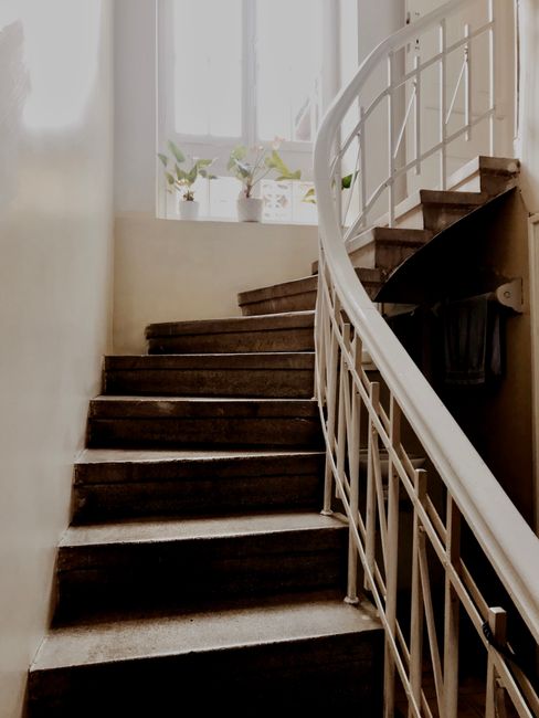 The image depicts a narrow staircase inside a terraced home, with dark wooden steps and a white curved metal handrail on the right side. At the top of the stairs, a window allows natural light to illuminate the area, showing a windowsill with small potted plants. The walls surrounding the staircase are painted in a light, neutral color. The staircase appears to be part of a home undergoing a house removal or relocation process, with no movers or packing materials visible, but the setting suggests a typical interior landing within a residential property managed by Man with Van Welling for removals services.