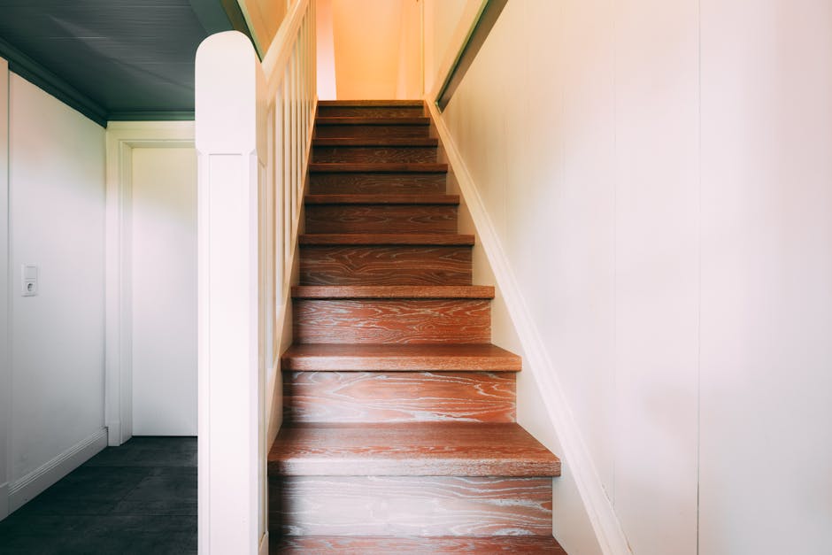 A woman with dark brown hair, wearing a beige coat, is carefully carrying a large cardboard box up a narrow staircase in a terraced home during a home relocation. Behind her, a man wearing a blue checkered shirt is also ascending the stairs, holding a similar box. The staircase features dark wooden steps with a black metal railing on the side, leading to an upper floor visible at the top. Natural light is coming through a window near the staircase, illuminating the scene. The concrete or tiled landing at the bottom of the stairs has some packing materials, and the interior walls are painted white. The image captures the process of furniture transport and packing during a move, with the two individuals working together to load boxes onto an upstairs landing, supported by a professional removal service from Man with Van Welling.