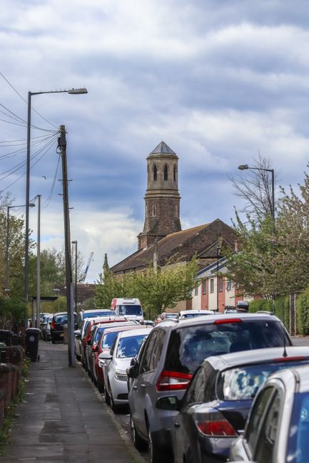 A street scene during daytime shows a row of parked cars along the pavement, including hatchbacks, sedans, and SUVs, with some vehicles partially visible. The pavement is narrow and bordered by a low fence and trees with light green foliage. In the background, there is a historic stone church with a tall bell tower, featuring a pyramidal roof and arched windows. Overhead, utility poles with power lines line the street, and a streetlamp is also visible on the left side. The sky above is partly cloudy with patches of blue. This setting illustrates urban residential parking, which may require parking suspensions during home relocation or furniture transport, as facilitated by services like Man with Van Welling.