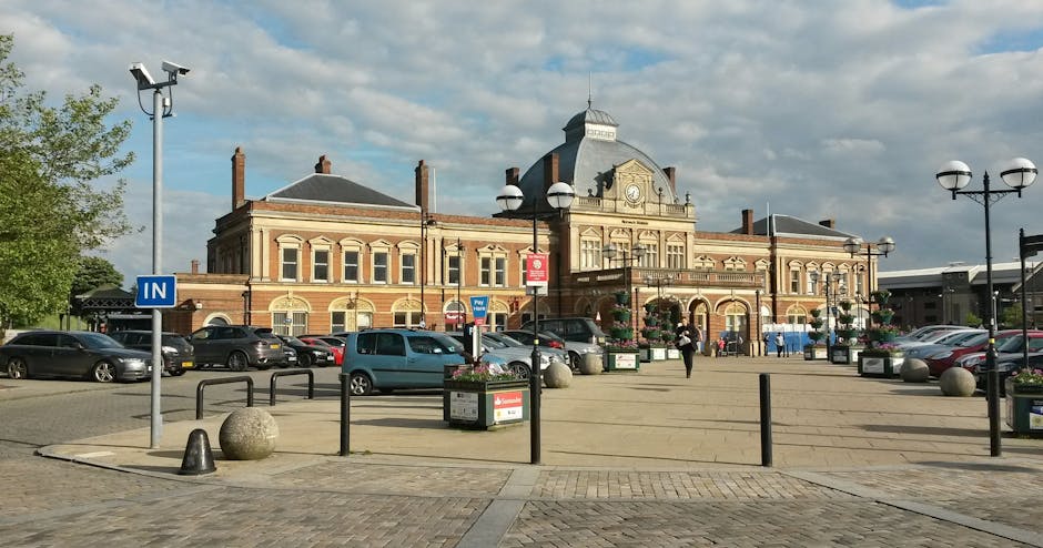 A large, historic brick building with a central clock tower and arched windows, situated in a town square with a paved open area in front. The square features parking spaces along the perimeter, with several parked cars, and is bordered by black metal barriers and large spherical concrete bollards. Decorative street lamps with multiple globes line the pedestrian area, and flower planters are placed near the barriers. In the foreground, a man is seen lifting a cardboard box wrapped in plastic, likely part of a home relocation or furniture transport process by Man with Van Welling. The scene is illuminated by daylight with a partly cloudy sky overhead, indicating an outdoor setting in a busy town environment suitable for moving services and parking suspensions when necessary for house removals in Welling.