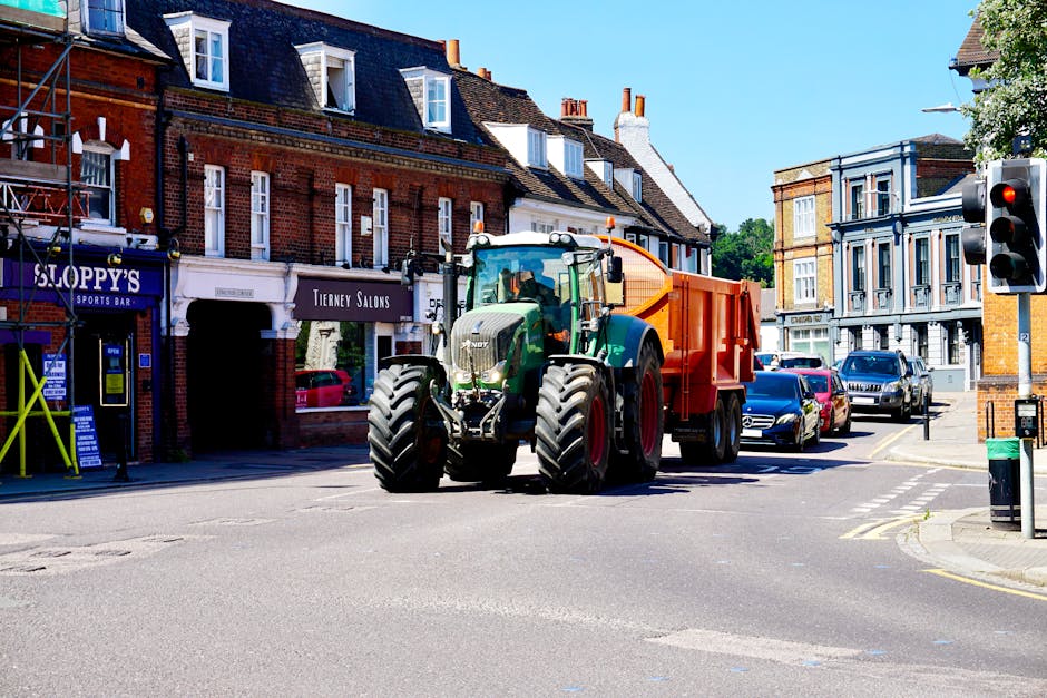 A large green and black tractor with a bright orange waste collection container attached to its rear is parked on a residential street in Welling. The tractor is situated at an intersection with a pedestrian crossing on the asphalt road, surrounded by parked cars on both sides. The scene takes place during daytime with clear blue skies, and traditional brick buildings with white-framed windows and signs for local shops such as 'Sloppy's Sports Bar' and 'Tierney Salons' are visible in the background. The tractor, which appears to be part of home relocation or waste disposal services, is positioned close to a traffic light and a recycling bin on the pavement, illustrating equipment used in moving or waste collection process as associated with house removals and clearance tasks, as detailed on manwithvanwelling.co.uk.