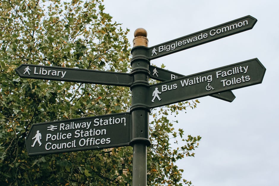 A black directional signpost with six arms pointing in different directions, situated outdoors against a background of leafy trees and an overcast sky. The signpost features white text indicating nearby locations including 'Biggleswade Common,' 'Library,' 'Bus Waiting Facility,' 'Toilets,' 'Railway Station,' 'Police Station,' and 'Council Offices.' The signs have pedestrian icons and wheelchair symbols, with some signs showing walking figures, indicating accessible routes. The signpost is anchored on a metal pole, which is visible in the center of the image. This scene relates to local navigation and can be associated with household relocation or community moves, which may involve planning routes with local amenities during moving services offered by Man with Van Welling, providing support for house removals and furniture transport in the Welling area.