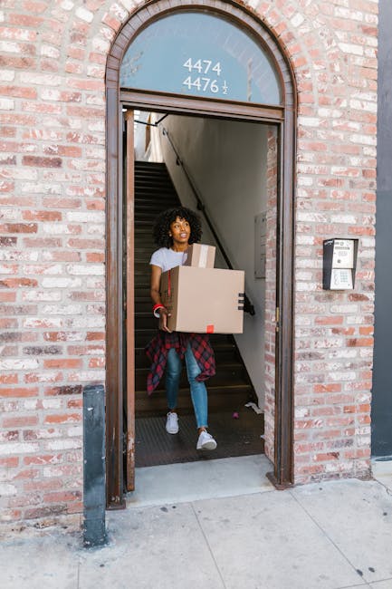 A woman with curly hair, dressed in casual clothing including a white t-shirt and jeans, is standing at the entrance of a brick building, carrying a cardboard box in her arms. Behind her, the interior staircase is visible, suggesting she is engaged in a home relocation process. She is wearing white sneakers, and her expression appears focused. In the background, part of a doorframe and external building facade are visible, with an intercom system mounted beside the entrance. The setting indicates an outdoor area adjacent to a residential or commercial property, with the woman likely involved in furniture transport or packing during a removal service, as offered by Man with Van Welling. The image emphasizes the loading and moving procedures associated with house removals in the Welling area, aligning with professional relocation and moving logistics.